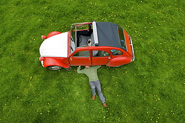 A man lying on his back under a red car, inspecting the underside of the car, Gloucestershire, England