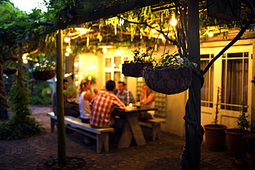 Group of people sitting outdoors at a table, hanging baskets in foreground, evening