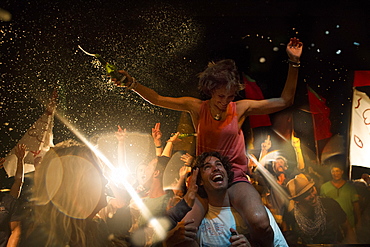 Revellers at an open air concert, smiling man carrying woman on his shoulders, arms outstretched, holding beer bottle