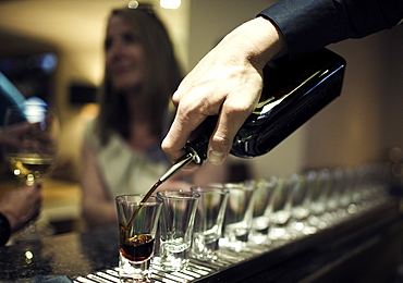 Close up of bartender pouring drinks from bottle into a row of shot glasses standing on bar counter, woman sitting in background