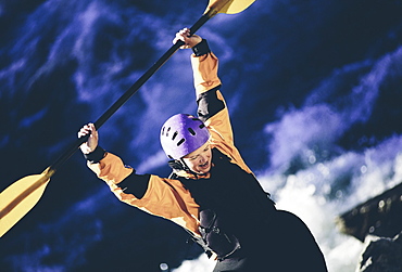 Female whitewater kayaker holding a paddle above her head river rapids in the background