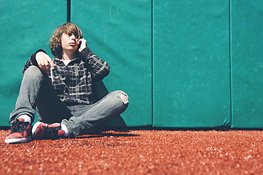 Teenage boy sitting against padded wall at sports field holding mobile phone