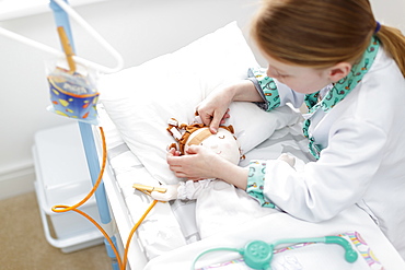 Young girl dressed as doctor putting plaster on doll's head in make-believe hospital bed