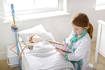 Young girl dressed as doctor writing notes next to pretend patient in make-believe hospital bed