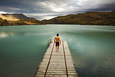 A young man walking down a wooden pier, towards calm lake surrounded by mountains in Torres del Paine National Park, Chile, Torres del Paine National Park, Chile.