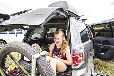 Teenage girl sitting on the tailgate of an SUV looking at vista, New Mexico, United States