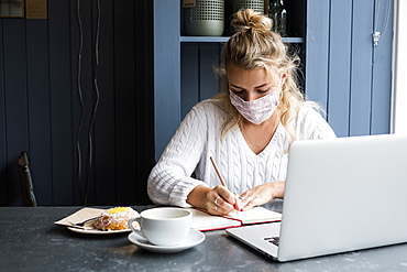 Woman wearing face mask sitting alone with a laptop, writing in note book, working remotely, Watlington, Oxfordshire, United Kingdom