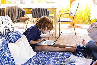 Boy with brown hair sitting on outdoor bed, doing homework on laptop, New Mexico, United States