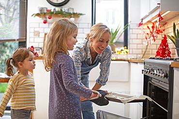 Blond woman wearing blue apron and two girls standing in kitchen, baking Christmas cookies