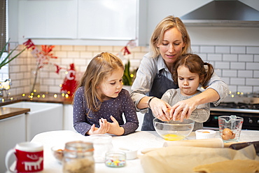 Blond woman wearing blue apron and two girls standing in kitchen, baking Christmas cookies