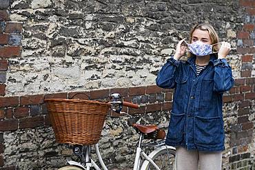 Young blond woman standing next to bicycle with basket, putting on face mask, Oxfordshire, England, United Kingdom