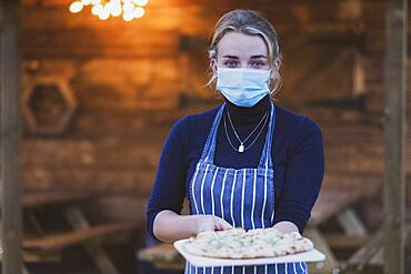 Woman waitress in apron and face mask holding plate of pizza