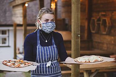 Woman waitress in apron and face mask holding plate of pizza