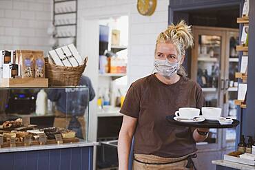 Waitress wearing face mask working in a cafe, carrying tray with coffee cups