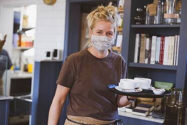 Waitress wearing face mask working in a cafe, carrying tray with coffee cups