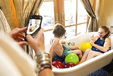 Person taking a smart phone photograph of a young boy and his older sister in bathtub with water balloons