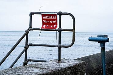 Red and white Covid-19 distancing sign on a harbour wall railing.