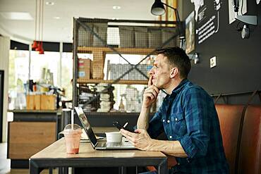 Man seated in a cafe using a laptop, wearing headphones, taking an online call.