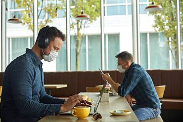 Two men seated in a cafe, working remotely using a  laptop and smart phone