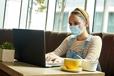 Woman wearing a face mask seated in a cafe using a laptop