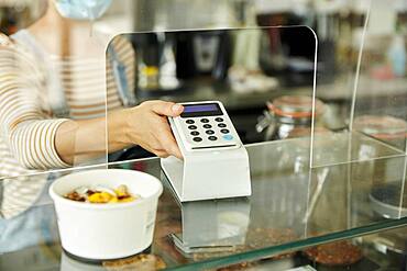 Woman in a face mask behind cafe safety screen, offering a contactless payment terminal to customer