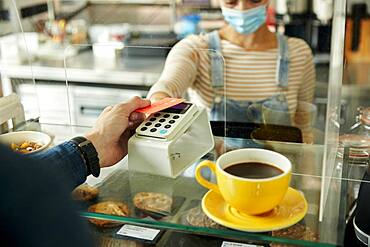 Woman in a face mask behind cafe safety screen, offering a contactless payment terminal to customer