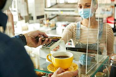 Woman in a face mask behind cafe safety screen, offering a contactless payment terminal to customer