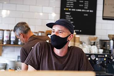 Male barista wearing black baseball cap and face mask working behind counter.