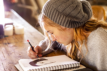 Teenage girl in a woolly hat drawing with a pencil on a sketchpad.