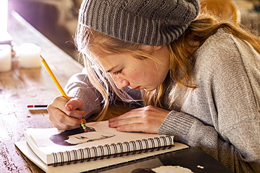 Teenage girl in a woolly hat drawing with a pencil on a sketchpad.