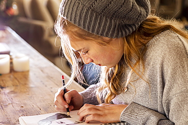 Teenage girl in a woolly hat drawing with a pencil on a sketchpad.