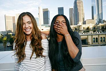 Two young women on a rooftop posing for photographs, city skyline background.