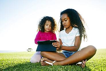 Young mixed race girl and her younger sister seated outdoors sharing a digital tablet