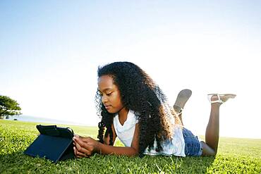 Nine year old mixed race girl lying on grass looking at a digital tablet.