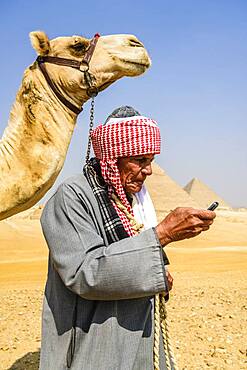 A guide in traditional Arab dress, robe and turban using a mobile phone, and his camel.