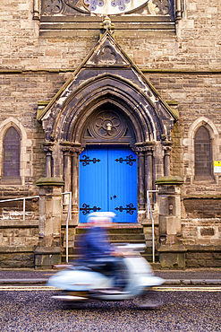 Person on a scooter driving past a gothic arch with bright blue door.