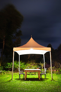 A small gazebo in a garden, a table and seat and lights at night