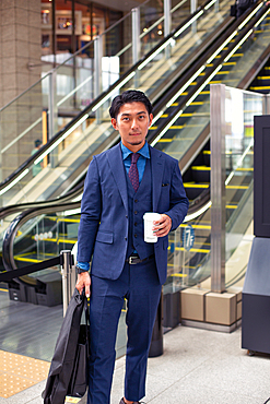 A young businessman in a blue suit on the move in a city downtown area, carrying a briefcase and cup of coffee.