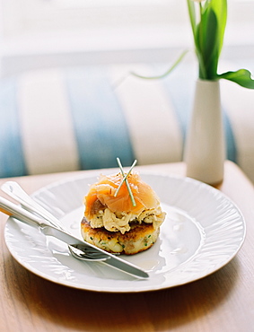 A plate with smoked salmon on a muffin, on a table. A vase with flowers and a striped sofa cushion, England