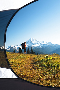 View from inside a camping tent of a man hiking across national forest land with Mount Baker in the distance, Cascades Range, Washington, USA