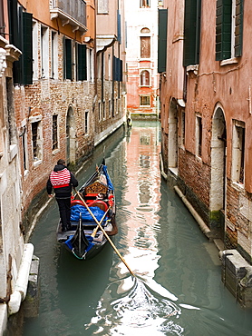 A gondola boat gliding down a small narrow waterway, between historic houses in the city of Venice, Venice, Italy