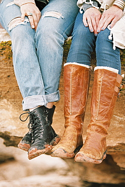 Close up of two women wearing leather boots sitting on a rock in a desert.