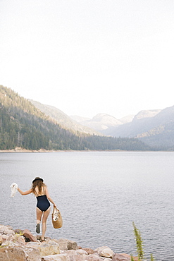 A woman in a swimsuit carrying a basket along the shore of a mountain lake.