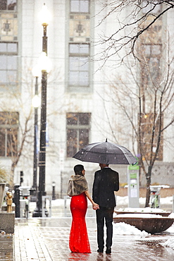 A woman in a long red evening dress with fishtail skirt and a fur stole, and a man in a suit, walking through snow in the city.