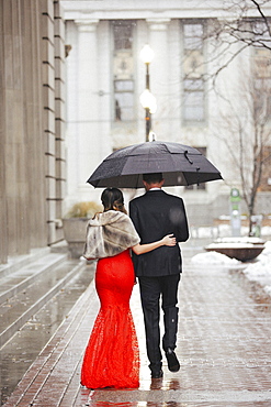 A woman in a long red evening dress with fishtail skirt and a fur stole, and a man in a suit, walking through a city.