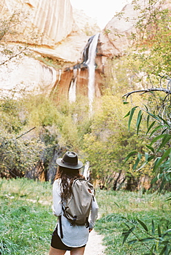 A woman carrying a backpack, walking under a tall cliff in a canyon. Waterfall, United States of America