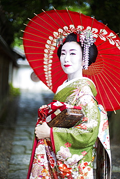 A woman dressed in the traditional geisha style, wearing a kimono and obi, with an elaborate hairstyle and floral hair clips, with white face makeup with bright red lips and dark eyes holding a red paper parasol, Japan