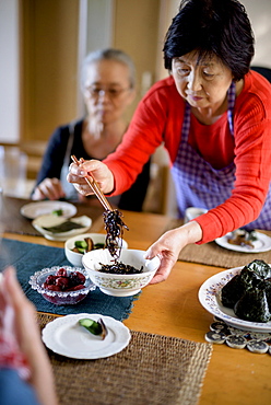 Woman wearing apron standing at kitchen table, holding bowl, serving food with chopsticks, Japan