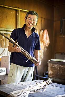 Japanese man standing in a traditional Washi workshop, holding a bunch of mulberry twigs, plant fibre ingredients and strips of fibre on a table, Kyushu, Japan