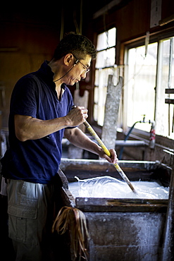 Japanese man stirring a vat of liquid in a workshop with a stick, making traditional Washi paper, Kyushu, Japan